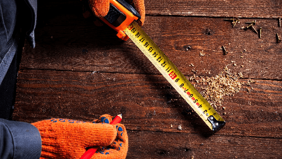 Gloved hands marking a measurement on a wooden board with a tape measure and pencil.