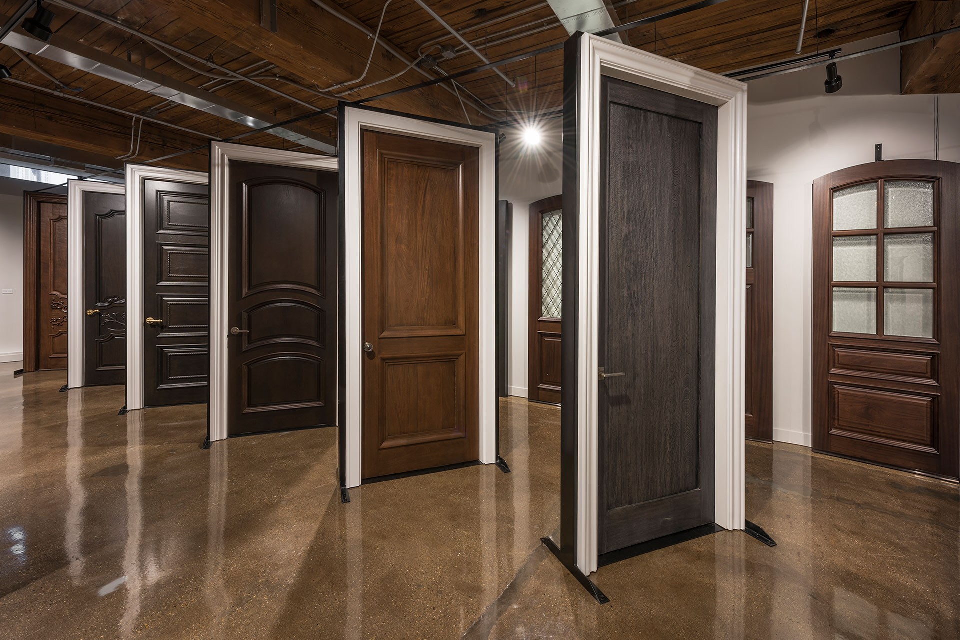 A door showroom with a dozen hardwood doors mounted upright on stands, each in a different panel and glazing style.