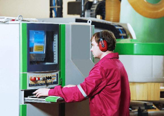 A machinist in ear defenders working at a CNC control panel on the shop floor.
