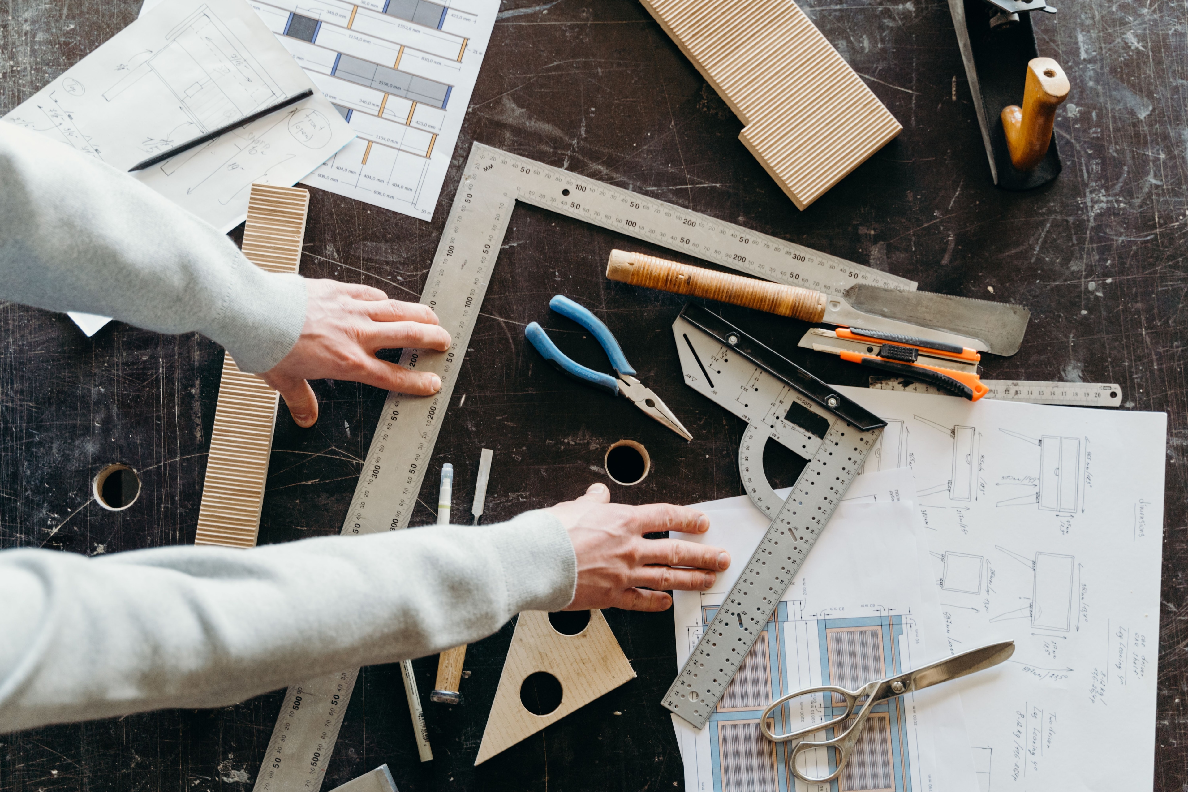 Overhead view of a workbench with two hands, a set square, rulers, a saw and hand-drawn door dimension drawings.