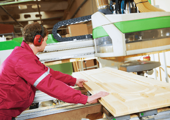 A shop-floor operator in ear defenders feeding a raised-panel door blank into a CNC machine.